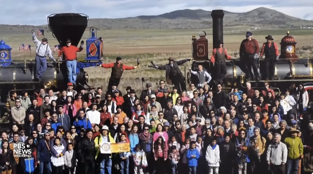 Large group of people posing in front of historic steam locomotives during a commemorative event, with mountains in the background. The event highlights the diverse community gathering to celebrate historical achievements and heritage.