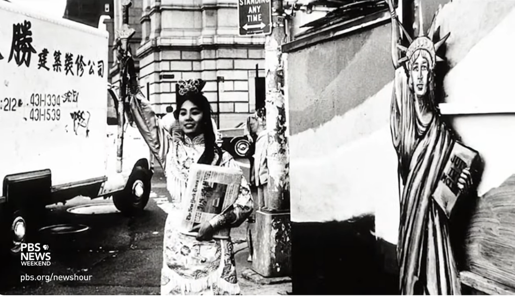 Black and white photo of a woman in traditional Asian attire holding a newspaper and a torch, standing next to a mural of the Statue of Liberty. The image captures a blend of cultural heritage and American symbolism in an urban setting.