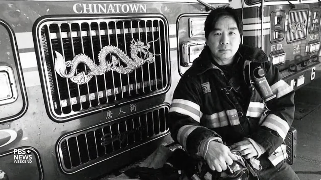 Black and white photo of an Asian American firefighter in uniform standing next to a fire truck with 'Chinatown' and a dragon emblem on the grille. The image highlights the presence and contribution of Asian Americans in the firefighting community. 