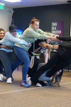 Group of people participating in a team-building exercise, pulling on each other in a line, with a sign in the background reading 'This is a trusted space.'