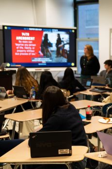 High school classroom with students using laptops, teacher presenting information on a large screen at the front.