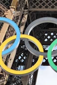The Olympic rings, colored blue, yellow, black, green, and red, are displayed prominently in front of the Eiffel Tower's iron lattice structure. The iconic rings symbolize the unity of the five inhabited continents. The Eiffel Tower's intricate metalwork is visible in the background, highlighting the location as Paris.