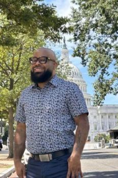 A man with glasses and a patterned shirt stands smiling in front of the U.S. Capitol building. The background includes trees and the dome of the Capitol, creating a pleasant and iconic outdoor scene.