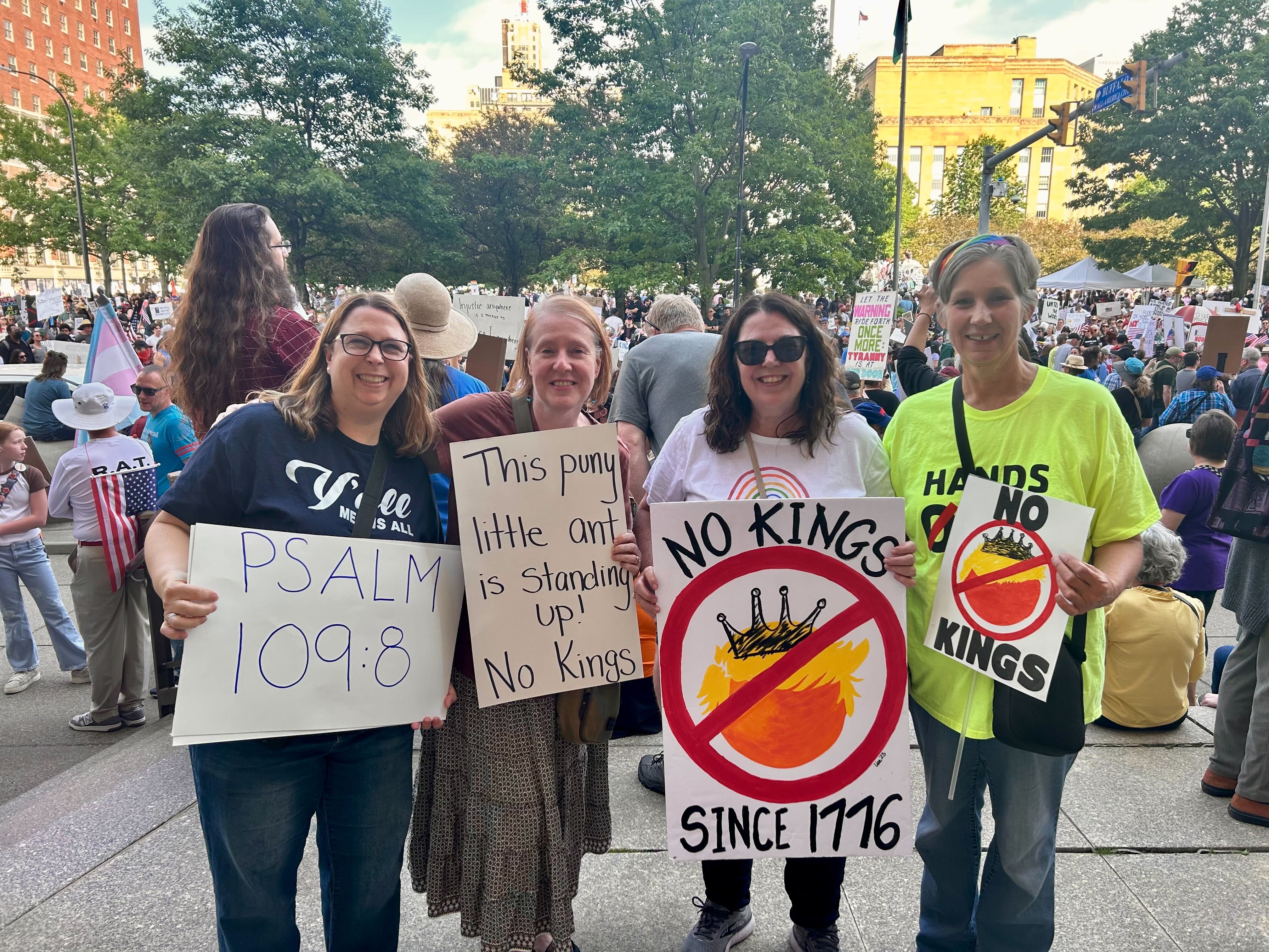 Four women stand together at a No Kings Day rally holding protest signs with messages such as “PSALM 109:8,” “This puny little ant is standing up!,” and “No Kings Since 1776,” featuring a crossed-out crown and orange hair. Behind them, a large, diverse crowd fills the public square, highlighting the energy, unity, and grassroots spirit described in Amber Chandler’s blog “No Kings Day: This Is What Democracy Looks Like.”