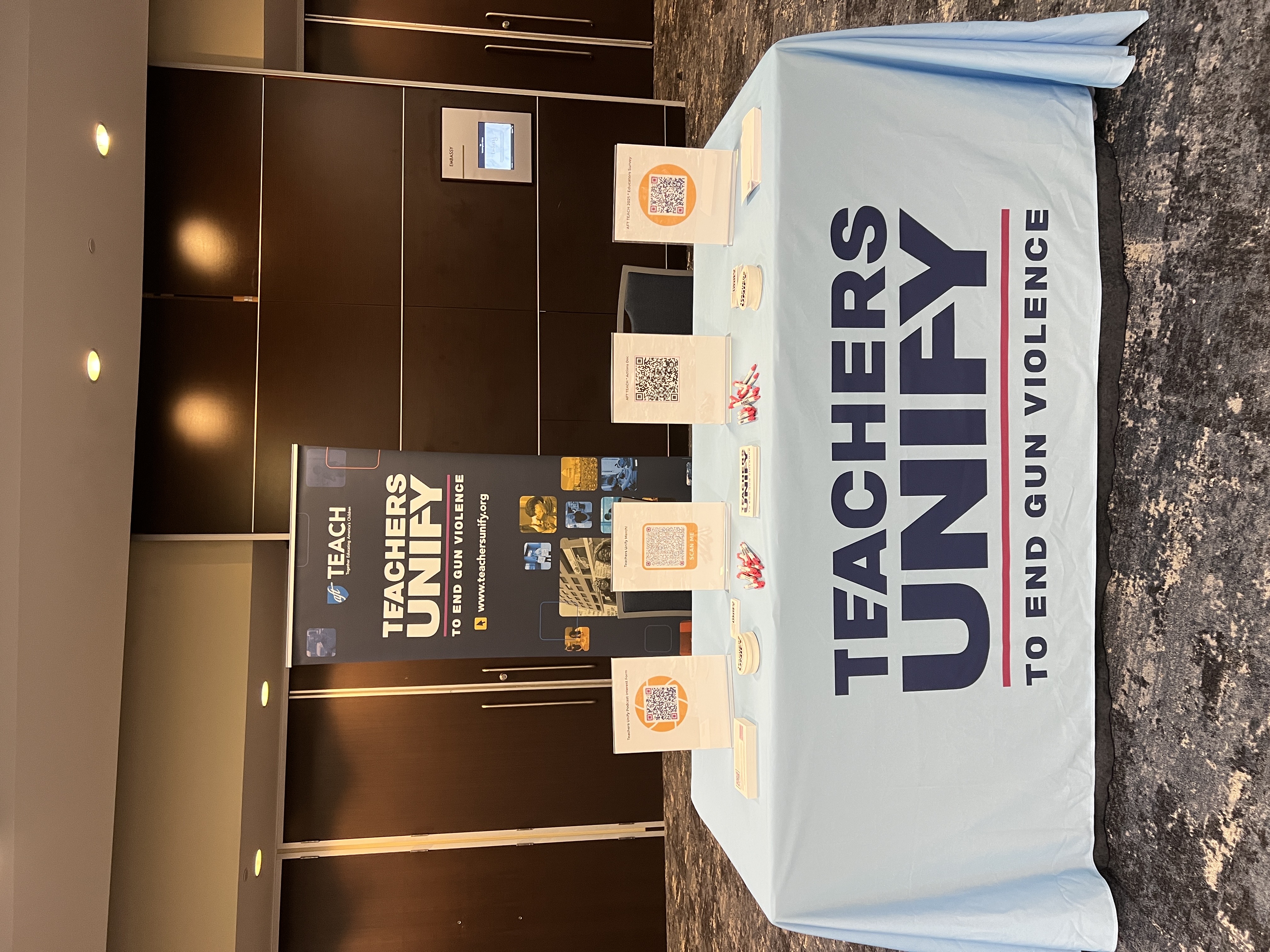 Teachers Unify to End Gun Violence table display at the AFT TEACH 2025 conference. The table is covered with a branded light blue tablecloth and features flyers, QR codes, buttons, pens, and informational materials. A vertical banner behind the table promotes the organization and its mission.