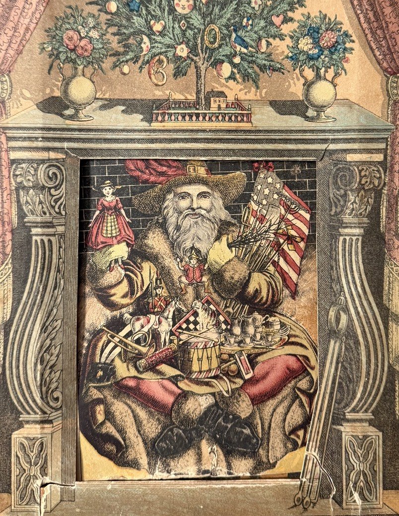 A richly illustrated 19th-century print of Santa Claus seated inside a fireplace, holding toys and American flags. He has a long white beard, a broad-brimmed hat with a red plume, and wears fur-trimmed robes. Santa is surrounded by dolls, drums, tea sets, and other gifts. Above him sits a decorated Christmas tree on the mantel with faces, ornaments, and a miniature house, flanked by floral vases. The style reflects patriotic American themes and Victorian-era holiday traditions.
